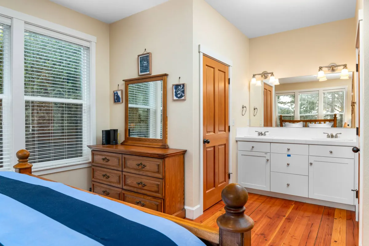 A tranquil bedroom with blue bedding, a wooden dresser topped with a large mirror, dual sink vanity, and gentle sunlight streaming in. The space is serene and ideal for preparing for the day or unwinding in comfort.