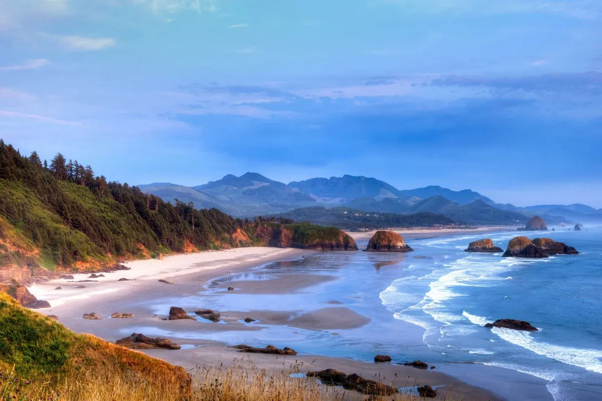 This image captures a sweeping coastal view with ocean waves rolling onto the beach, dramatic rock formations, distant mountains, and a broad shoreline. The natural beauty is on full display, inviting exploration and outdoor adventure.