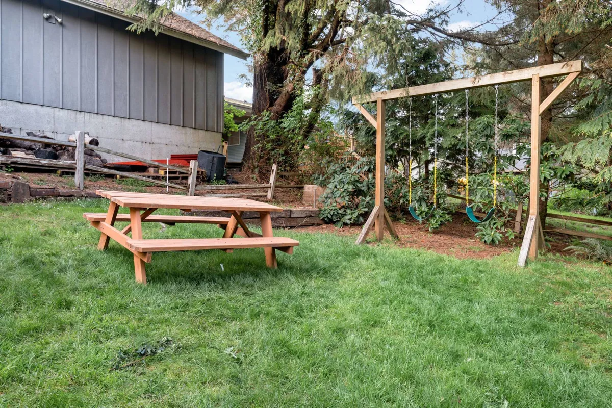 This image showcases a vibrant backyard scene with a picnic table set among lush greenery and a swing set nearby. The area is bathed in soft natural light, creating an inviting and playful atmosphere. The space is clearly designed for both relaxation and family fun, with ample room for group activities or quiet outdoor moments.
