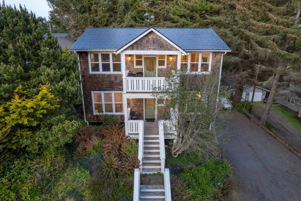The image showcases the striking wooden exterior of the Pacific Star home, framed by lush greenery and mature trees. A staircase leads up to the inviting entrance, while the surrounding garden hints at outdoor spaces perfect for relaxation and family gatherings. The tranquil setting suggests opportunities for enjoying nature, from stargazing on the porch to morning yoga on the lawn.