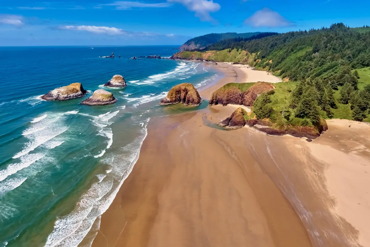 An expansive view of the sandy beach framed by rock formations, gentle waves, and a lush green hillside. The image evokes a sense of exploration and discovery, inviting visitors to walk along the shore, search for shells, or simply relax by the tide pools.
