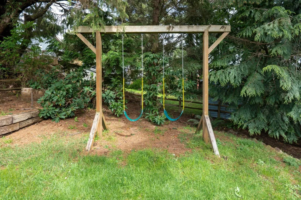 A joyful backyard setting with a swing set standing on a soft grassy area, surrounded by lush greenery. Children can be imagined playing here, and the space feels open and safe for family gatherings and outdoor fun. The mood is cheerful and nostalgic, evoking playful memories and carefree afternoons.