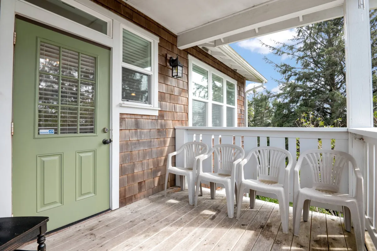 A relaxed exterior scene featuring a green door, wooden siding, and a few plastic chairs, suggesting a casual spot for guests to gather and enjoy the peaceful surroundings. The ambiance hints at tranquil moments and sociable evenings.