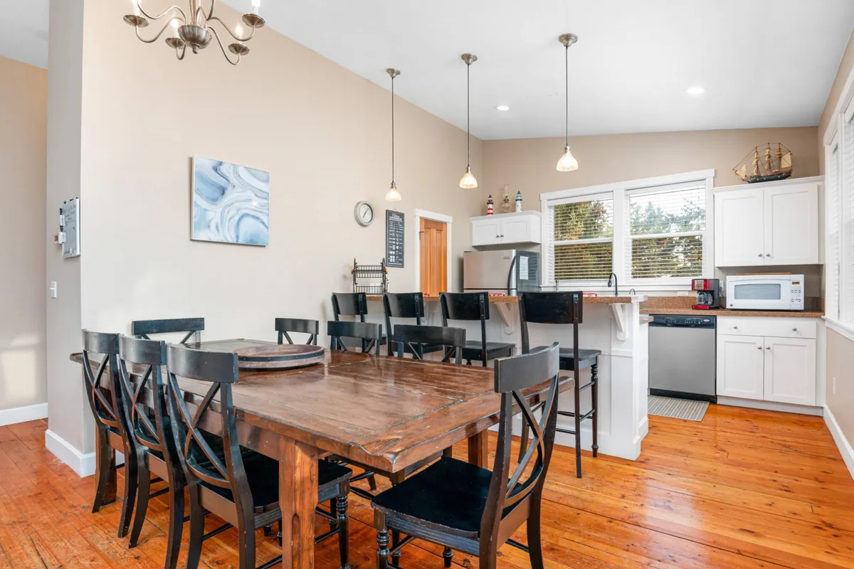 The image displays a large dining table set adjacent to a bar counter and kitchen appliances, with artwork providing a touch of personality. The arrangement invites guests to come together for family meals, casual brunches, or festive dinner parties, all in a warm, communal atmosphere.