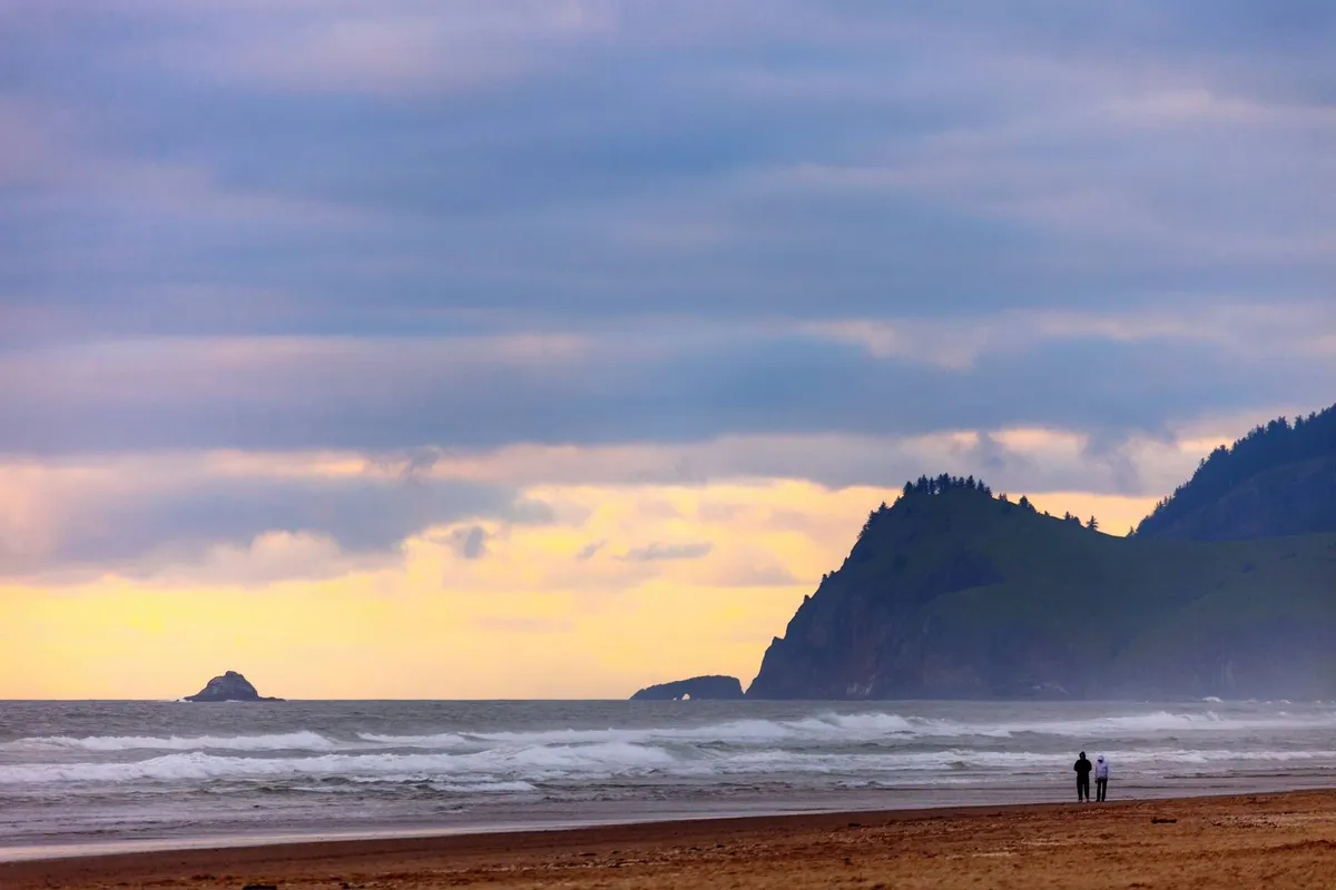 A romantic scene capturing a couple walking along the sandy beach at sunset, with ocean waves rolling in and distant cliffs providing a dramatic backdrop. The couple is collecting seashells, and the soft glow highlights the serene, natural beauty of the Oregon Coast.